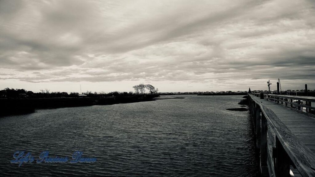 Black and white of a pier in Murrells Inlet trailing into the distance along the watershed. Cloudy storm clouds in the sky and a boat sits on the bank in the background.