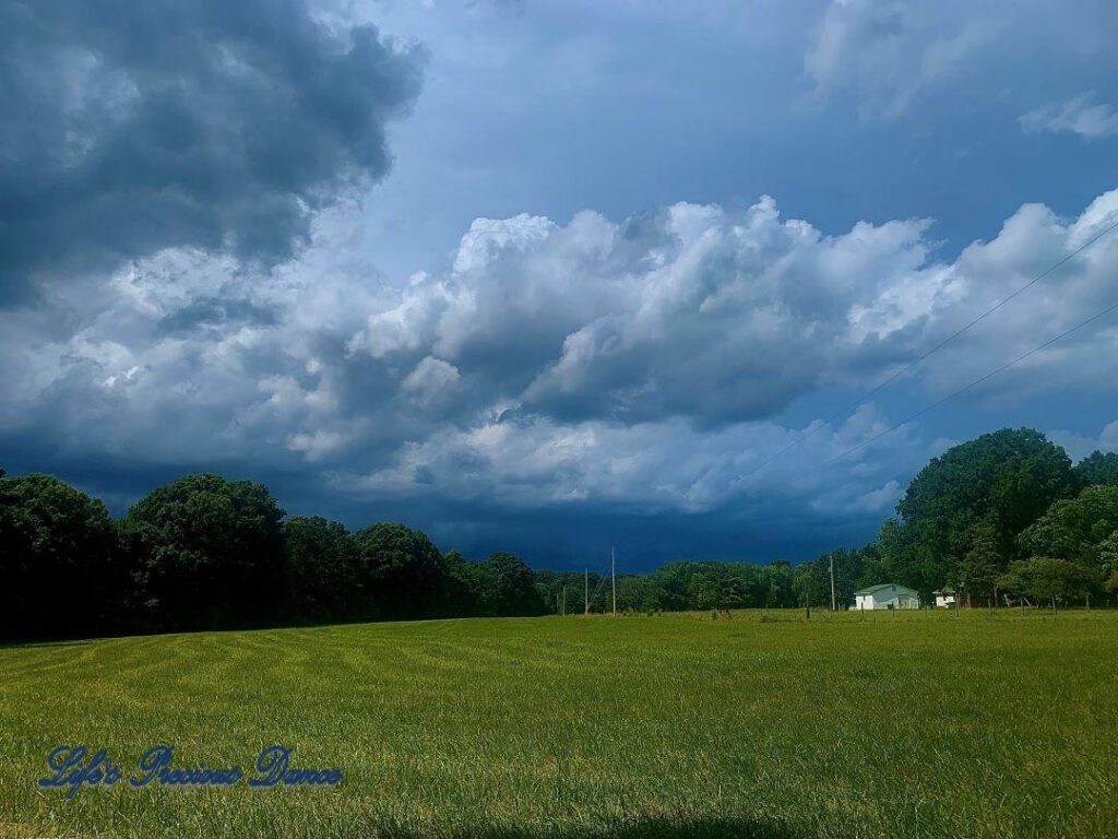 Ominous storm clouds rolling in above the trees, over a pasture. A farmhouse sits to the right.