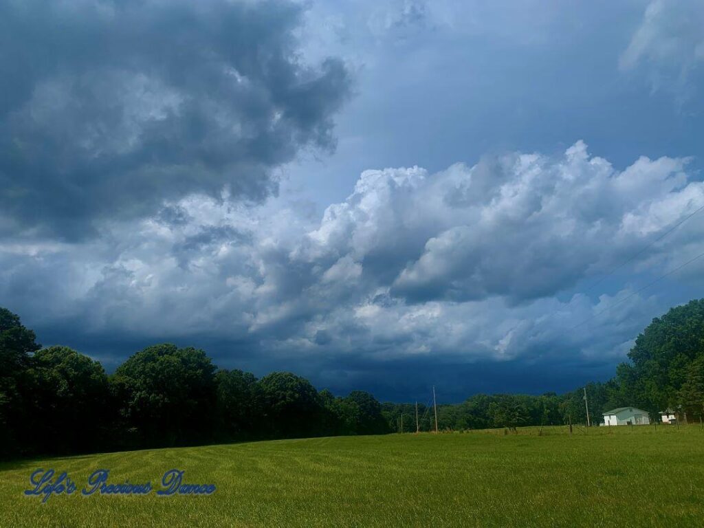 Ominous storm clouds rolling in above the trees, over a pasture. A farm house sits to the right.