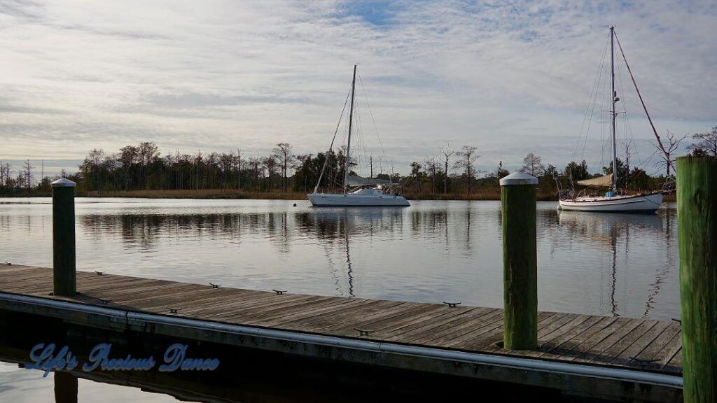 Sailboats resting in a harbor. A dock in the foreground and clouds overhead.