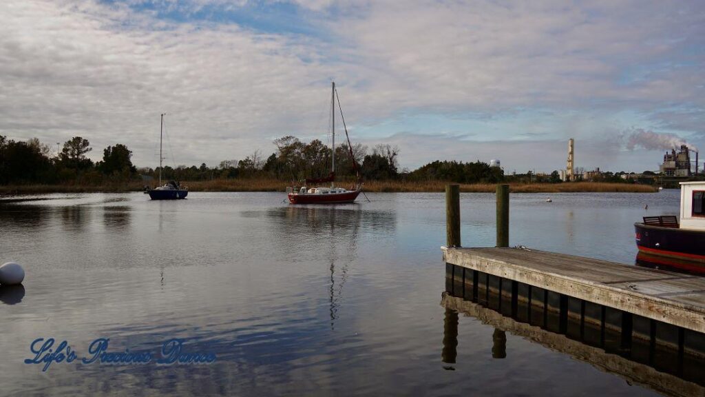 Sailboats resting in the harbor. A dock in the foreground, and clouds above, reflecting in the water.