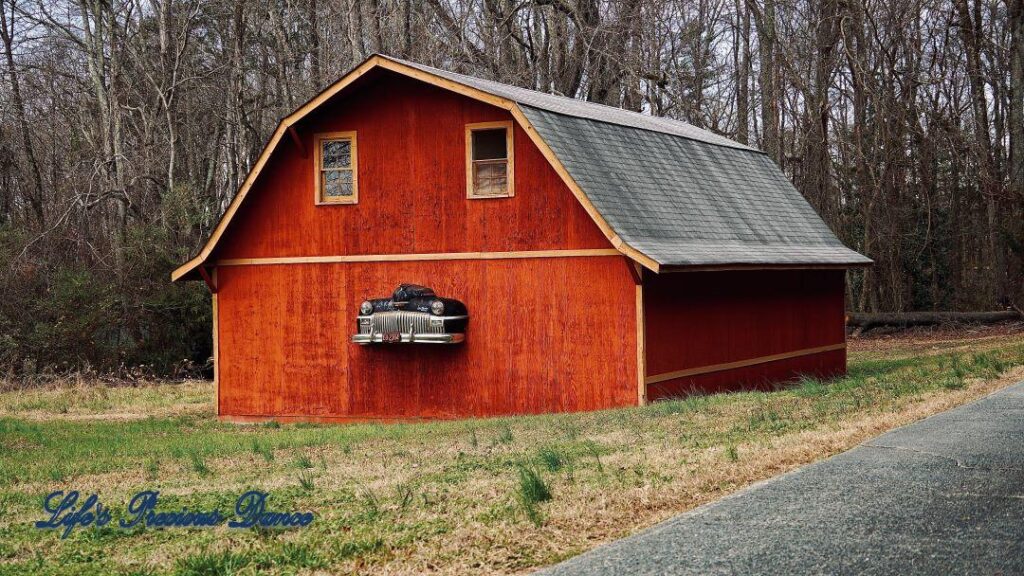 Rust colored barn with front end of classic DeSoto automobile hood protruding from structure.