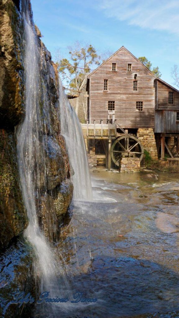 Close up of water cascading over a rock wall into creek. Yates MIll in the background.