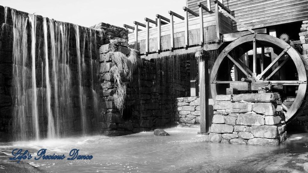 Black and white of pond water cascading over a rock wall beside an old mill.