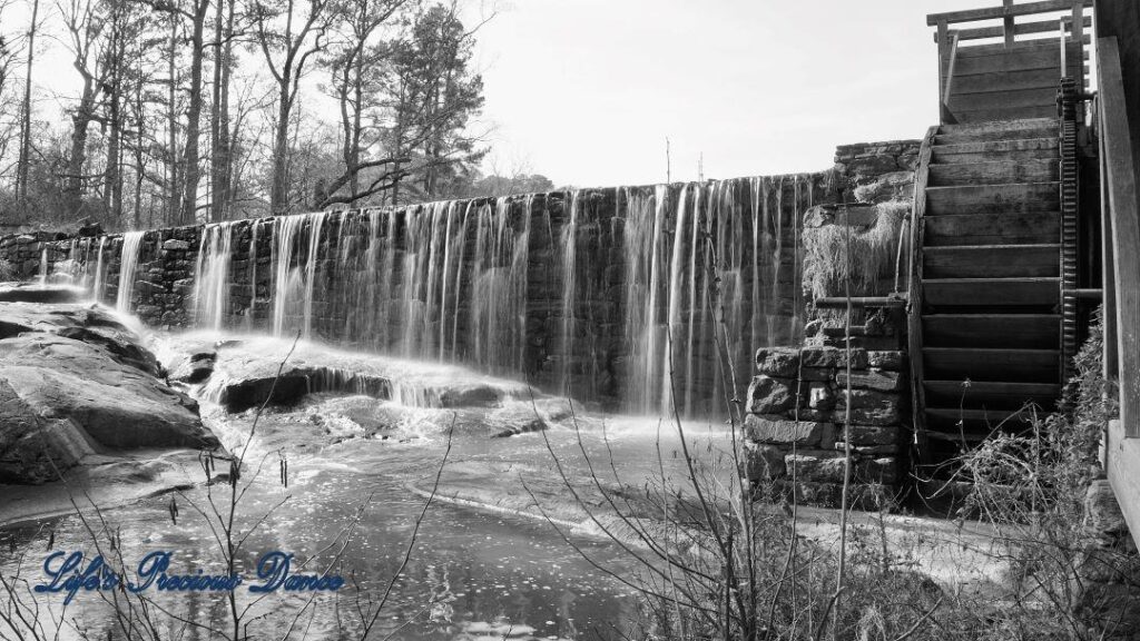 Black and white of water from pond cascading over rockwall into creek. Waterwheel of Yates MIll to the right.