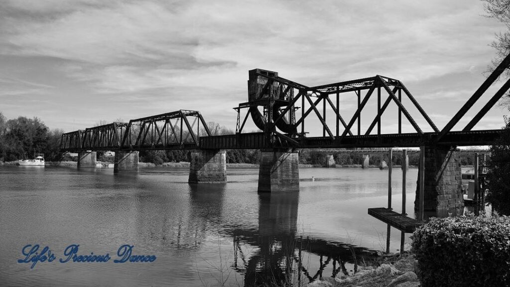 Black and white of a train trestle spanning the Savannah River. Clouds overhead and a boat in the background.