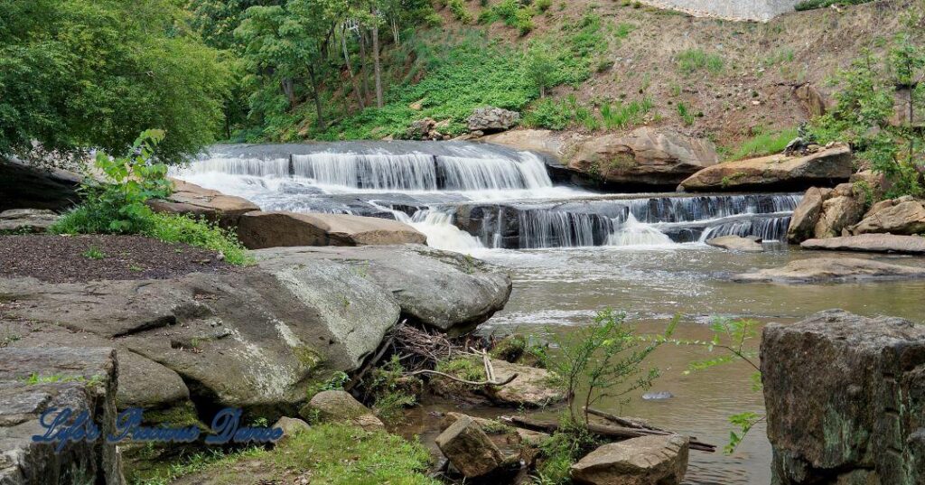 Lower section of Reedy Falls cascading over a rock ledge into the river. A metal sculpture sits on a boulder.