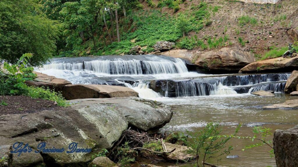 Lower section of Reedy Falls cascading over a rock ledge into the river. A metal sculpture sits on a boulder.