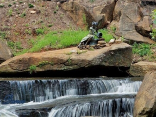 Lower section of Reedy Falls cascading over a rock ledge into the river. A metal sculpture sits on a boulder above.