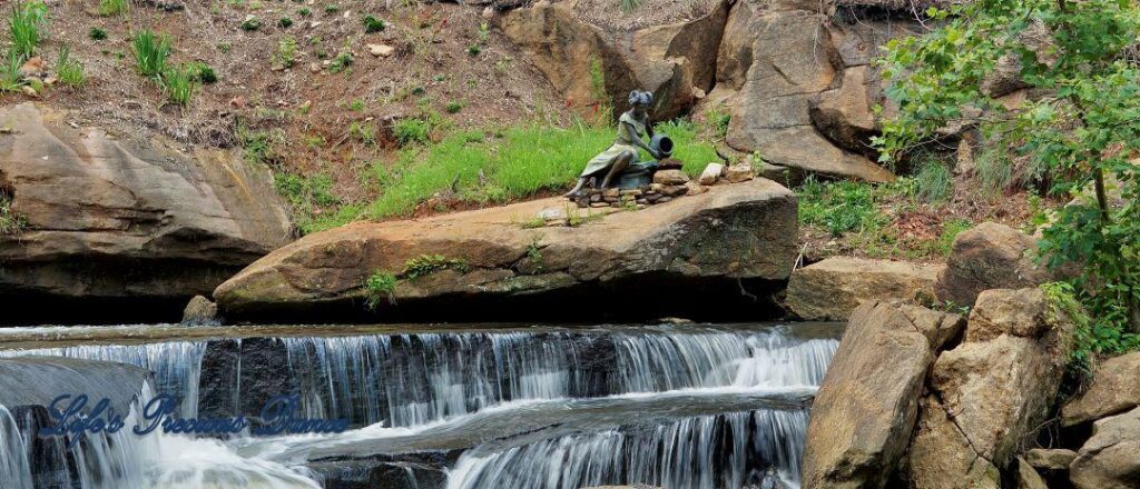 Lower section of Reedy Falls cascading over a rock ledge into the river. A metal sculpture sits on a boulder above.