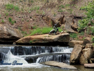 Lower section of Reedy Falls cascading over a rock ledge into the river. A metal sculpture sits on a boulder above.
