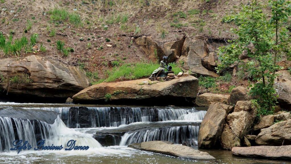 Lower section of Reedy Falls cascading over a rock ledge into the river. A metal sculpture sits on a boulder above.