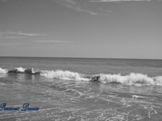 Black and white of waves crashing into the shore. A few passing clouds above.