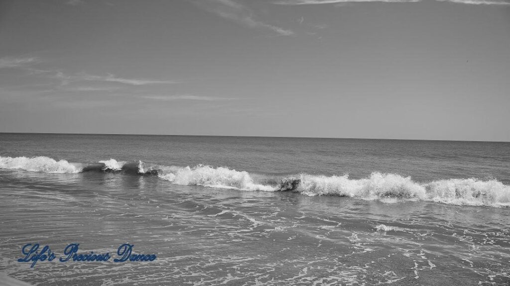 Black and white of waves crashing into the shore. A few passing clouds above.