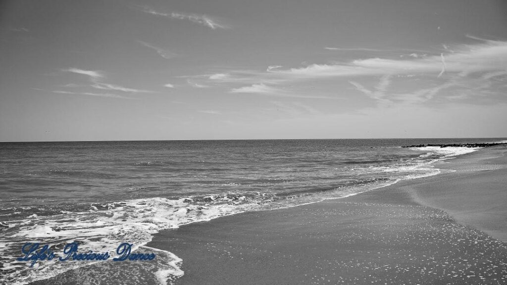 Black and white of waves rolling onto the shore. A few passing clouds above.