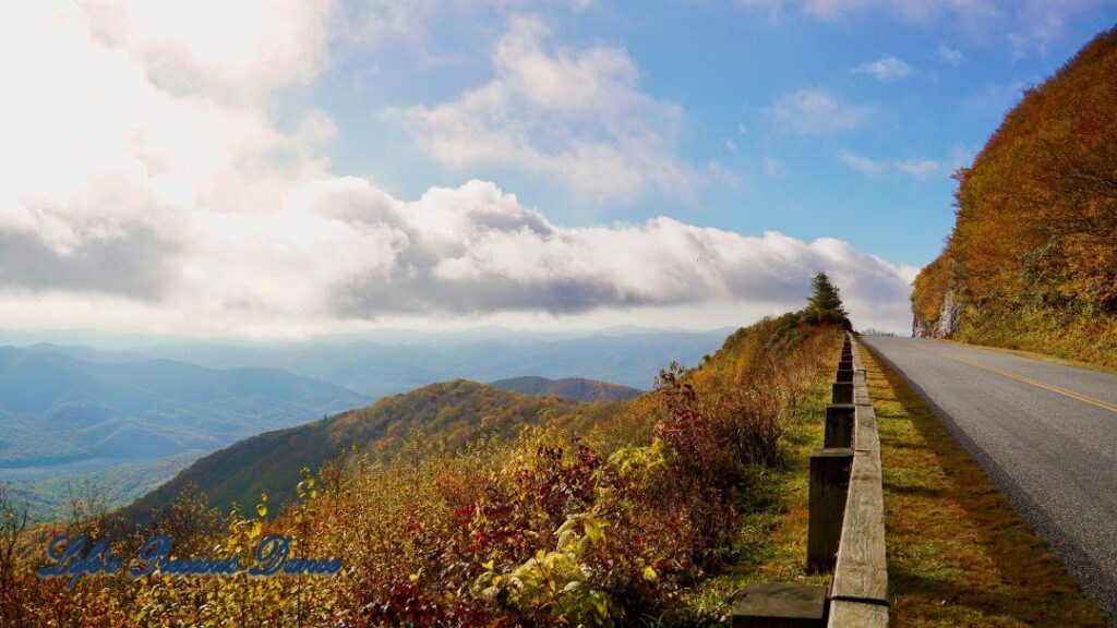 Landscape view of the colorful trees of the Blue Ridge Mountains. Passing clouds against a Carolina blue sky and the parkway to the right.