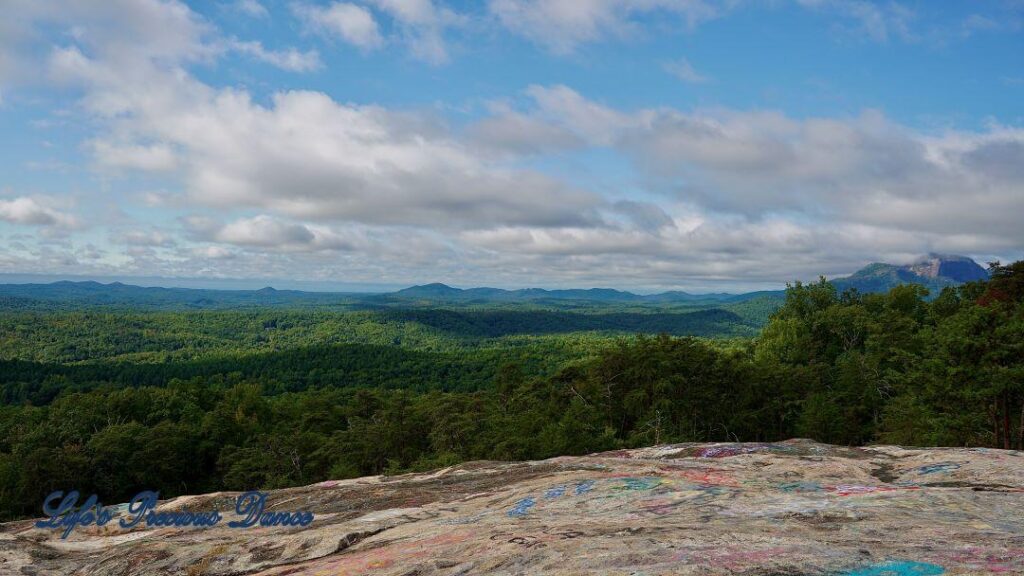 Landscape view overlooking a valley of trees. Clouds up above and mountains in the background.