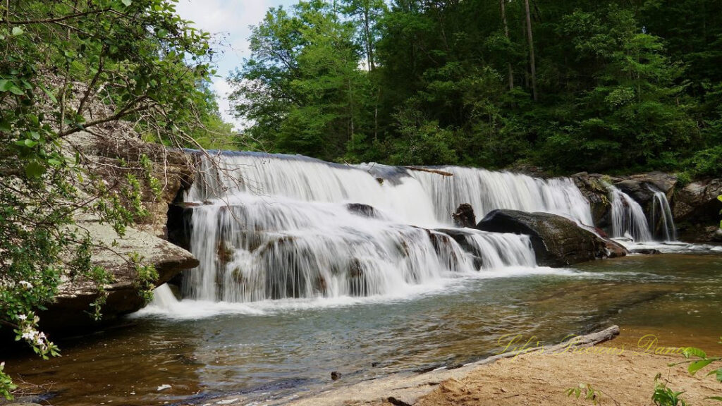 Riley Moore Falls spilling over rocks into the Chauga River. A sandy beach in the foreground.