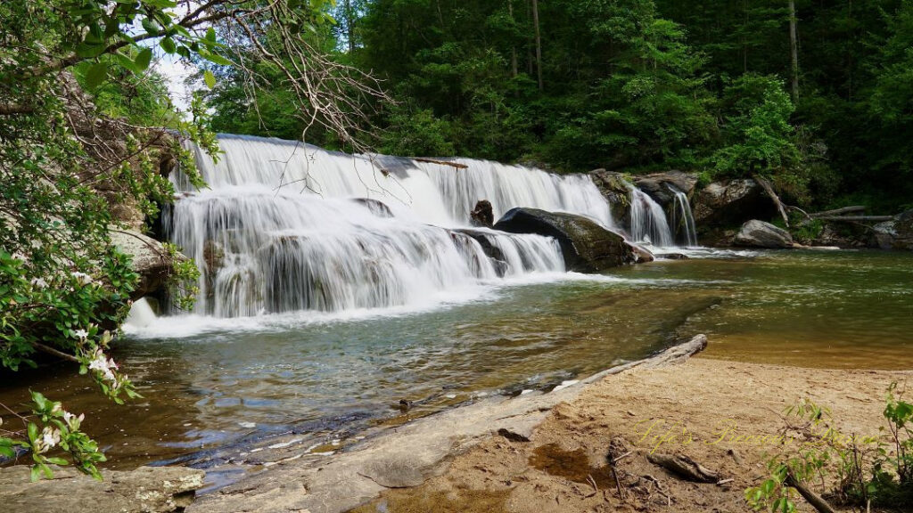 Riley Moore Falls spilling over rocks into the Chauga River. A sandy beach in the foreground.