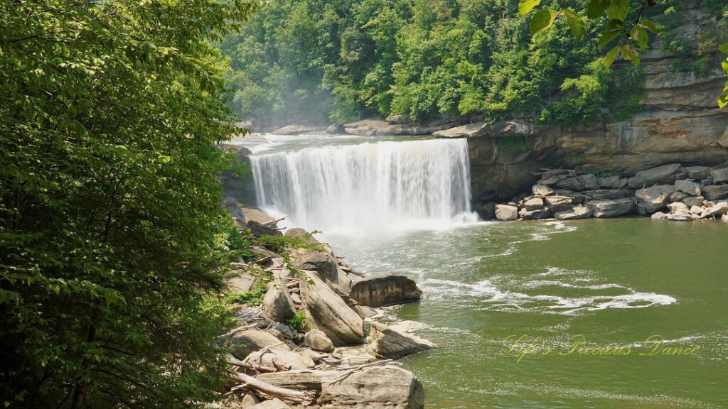 Cumberland Falls rapidly spilling into the river below. Trees and rockface mountains surrounding.