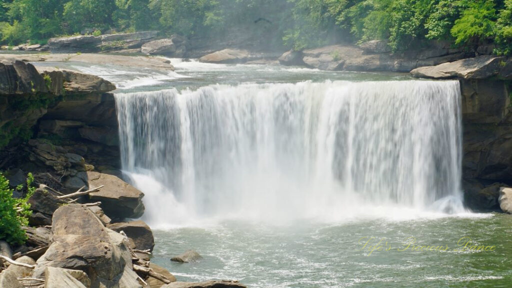 Cumberland Falls rapidly spilling into the river below. Trees and rockface mountains surrounding.