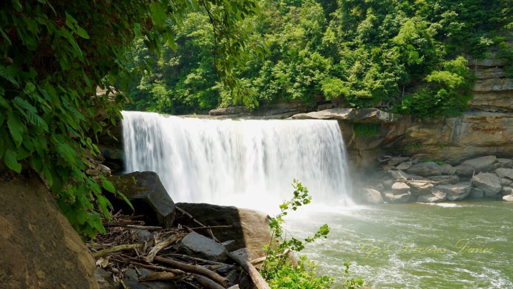 Cumberland Falls rapidly spilling into the river below. Trees and rockface mountains surrounding.