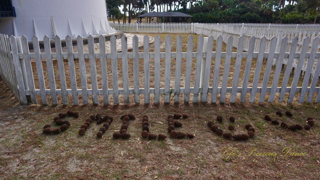 Pinecones spelling out &quot;Smile&quot; on the ground in front of a white picket fence at Hunting Island lighthouse.