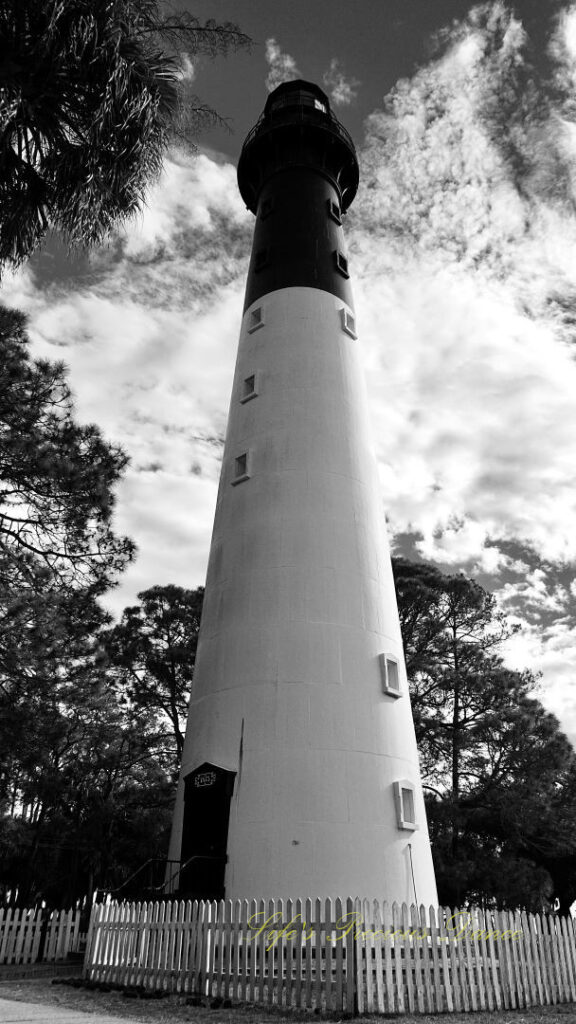 Black and white of the Hunting Island lighthouse against a mixed a blue sky with passing clouds. A white picket fence surrounding it.