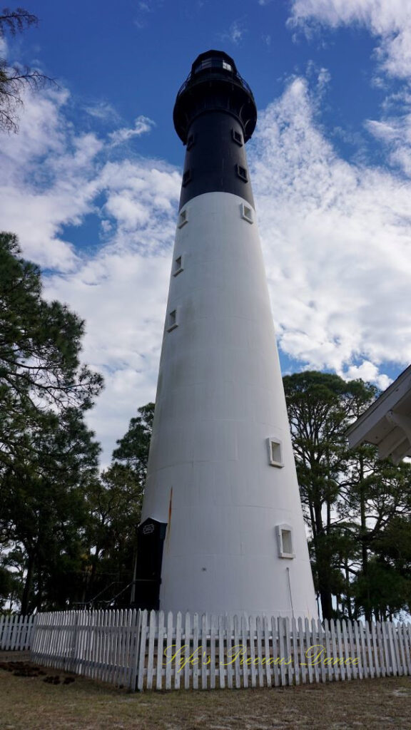 The Hunting Island lighthouse against a mixed a blue sky with passing clouds. A white picket fence surrounding it.