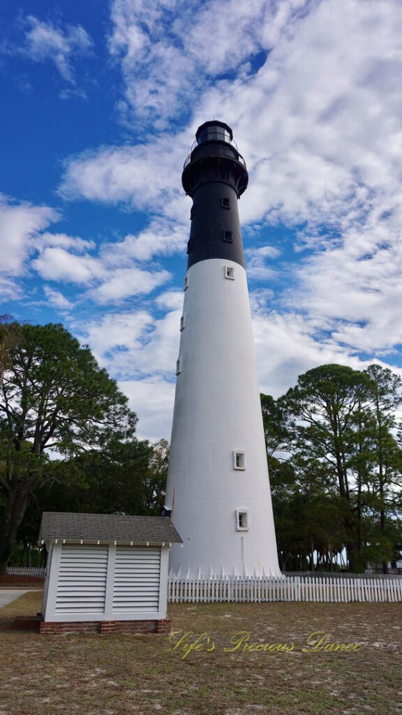 The Hunting Island lighthouse against a mixed a blue sky with passing clouds. A white picket fence surrounding it.