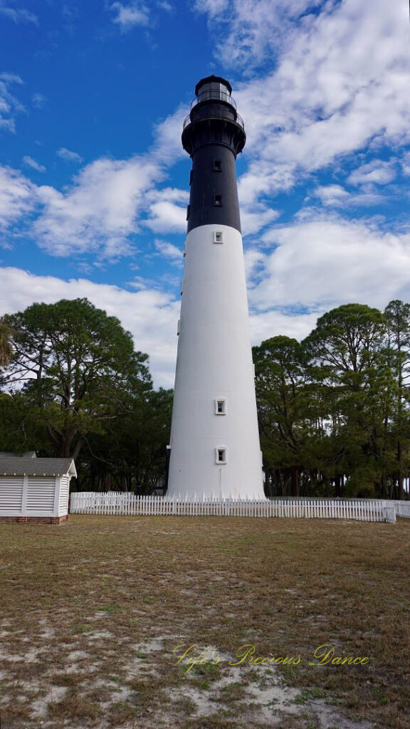 The Hunting Island lighthouse against a mixed a blue sky with passing clouds. A white picket fence surrounding it.