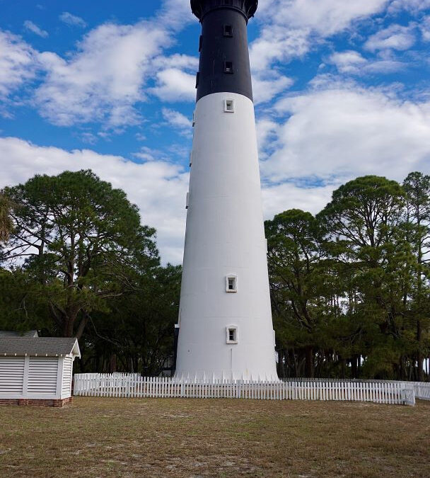 The Hunting Island lighthouse against a mixed a blue sky with passing clouds. A white picket fence surrounding it.