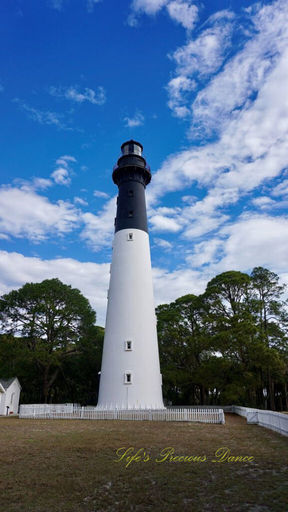 The Hunting Island lighthouse against a mixed a blue sky with passing clouds. A white picket fence surrounding it.