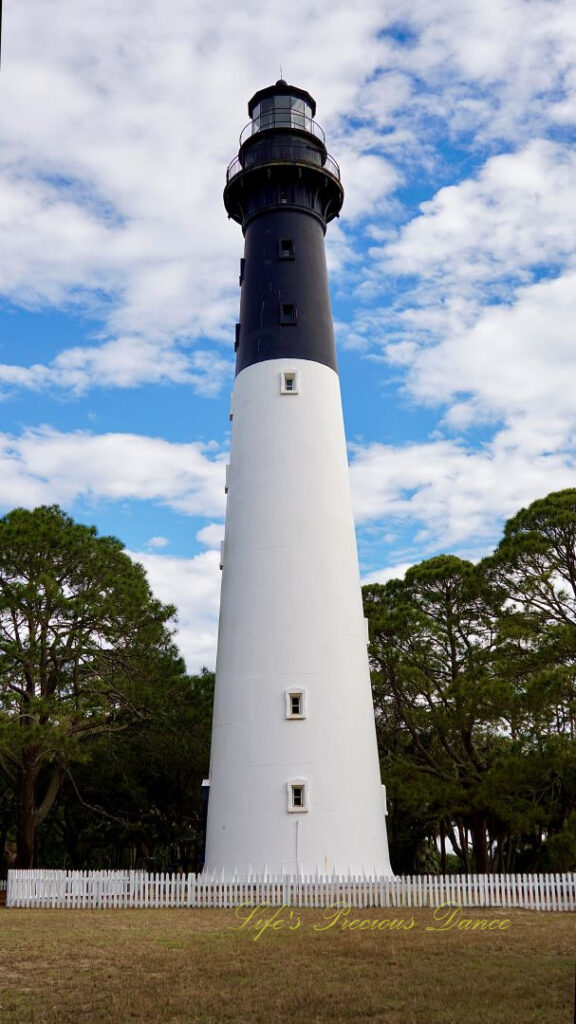 The Hunting Island lighthouse against a mixed a blue sky with passing clouds. A white picket fence surrounding it.