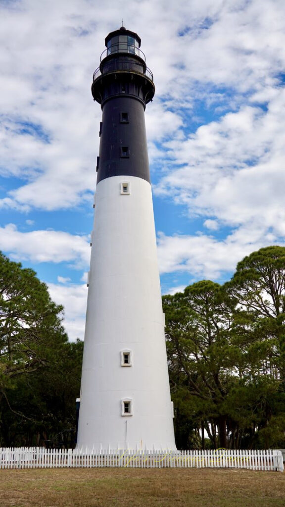 The Hunting Island lighthouse against a mixed a blue sky with passing clouds. A white picket fence surrounding it.