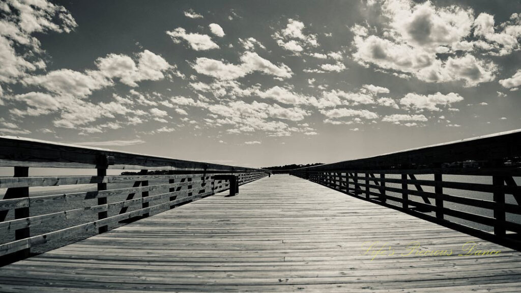 Black and white long view of a fishing pier at Hunting Island State Park. Fluffy clouds up above.