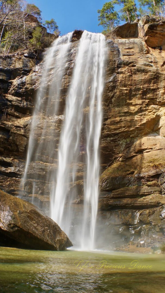 An 186 foot waterfall spilling over a rock face into a pool of water at Toccoa Falls. A large boulder glistens in the foreground.