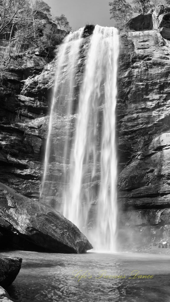 Black and white of an 186 foot waterfall spilling over a rock face into a pool of water at Toccoa Falls. A large boulder glistens in the foreground.