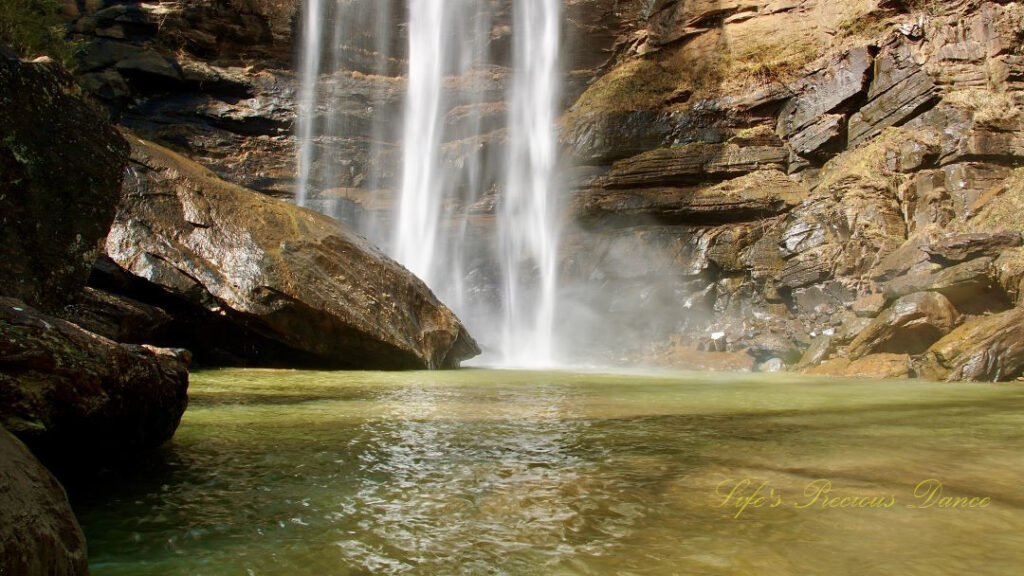 An 186 foot waterfall spilling over a rock face into a pool of water at Toccoa Falls. A large boulder glistens in the foreground.