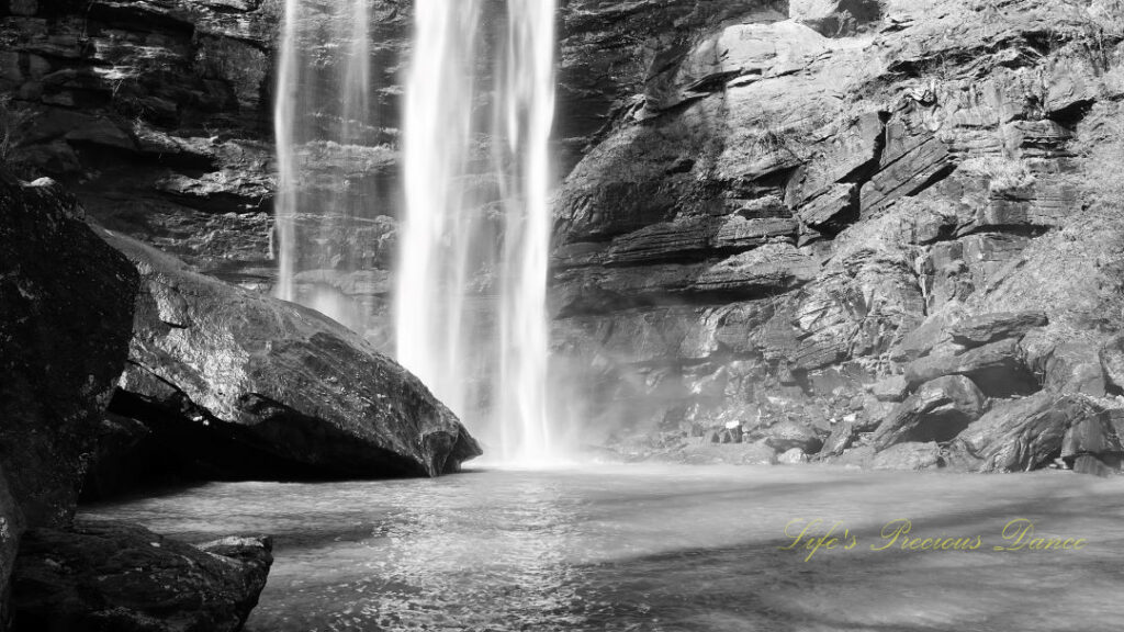 Black and white of a waterfall cascading down a rock face into a pool of water at Toccoa Falls. A large boulder glistening in the foreground.