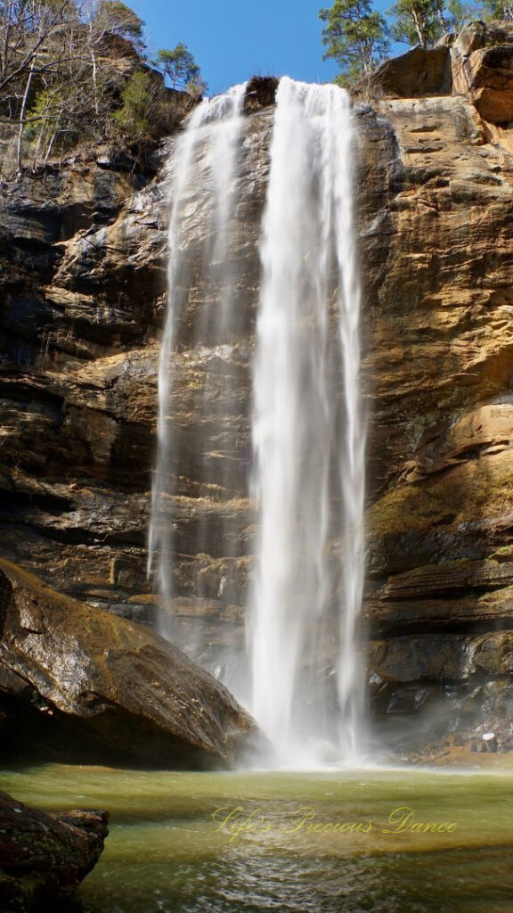 An 186 foot waterfall spilling over a rock face into a pool of water at Toccoa Falls. A large boulder glistening in the foreground.