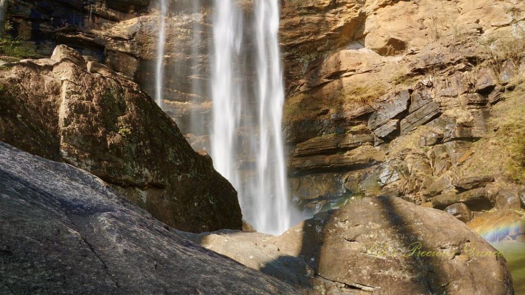 A rainbow reflecting in the mist above a pool of water as Toccoa Falls cascades down the rock face. Large boulders in the foreground.