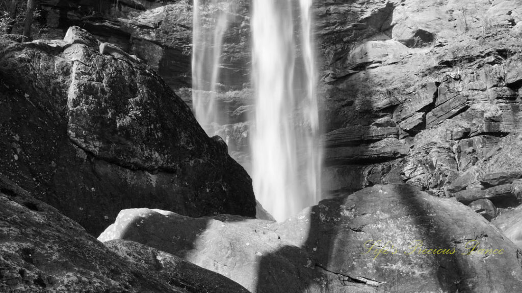 Black and white close up of a waterfall cascading down a rockface. Large boulders in the foreground.