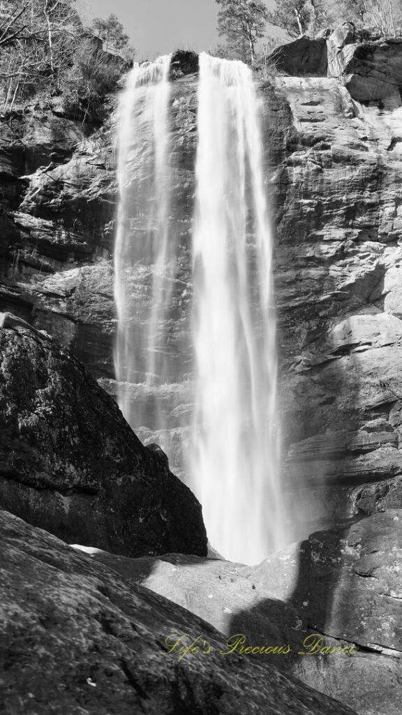 Black and white of an 186 foot waterfall spilling over a rock face into a pool of water at Toccoa Falls. A large boulder in the foreground.