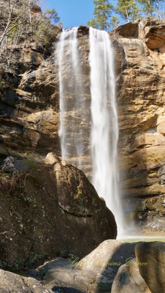 An 186 foot waterfall spilling over a rock face into a pool of water at Toccoa Falls. A large boulder in the foreground.