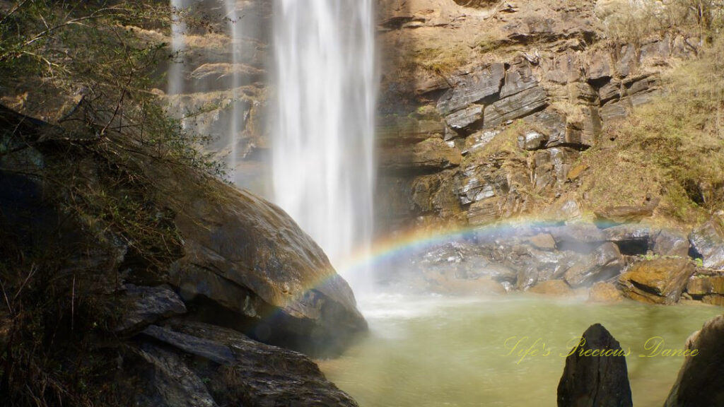 A rainbow reflecting in the mist above a pool of water as Toccoa Falls cascades down the rock face.