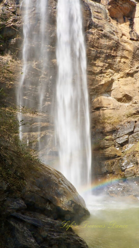 An 186 foot waterfall spilling over a rock face into a pool of water at Toccoa Falls. A rainbow is reflecting in the mist.