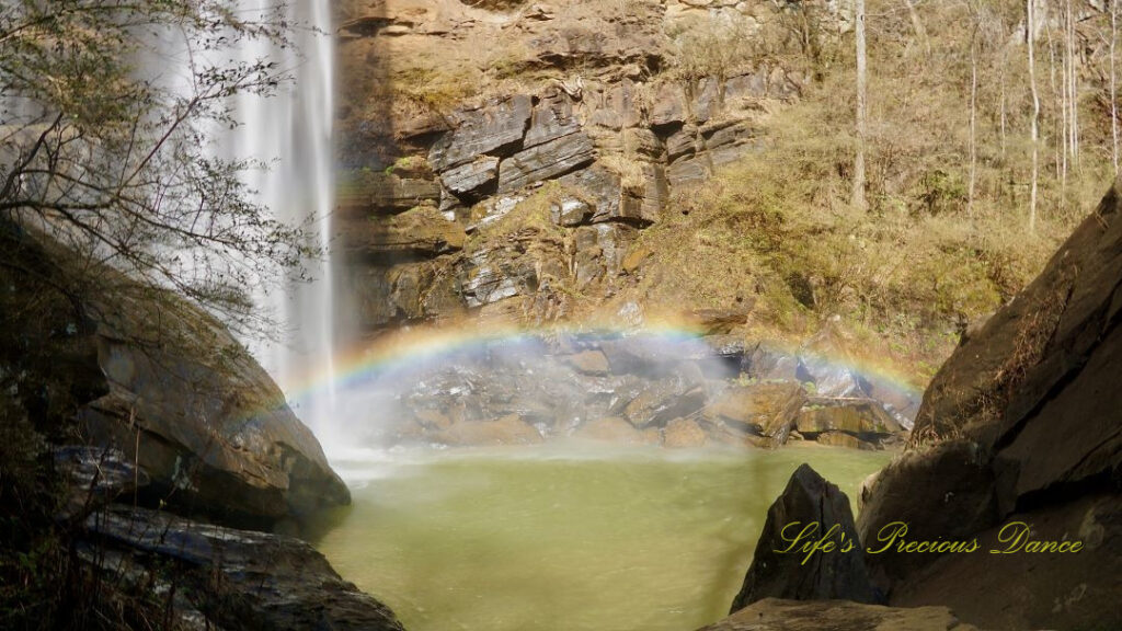 A rainbow reflecting in the mist above a pool of water as Toccoa Falls cascades down the rock face.