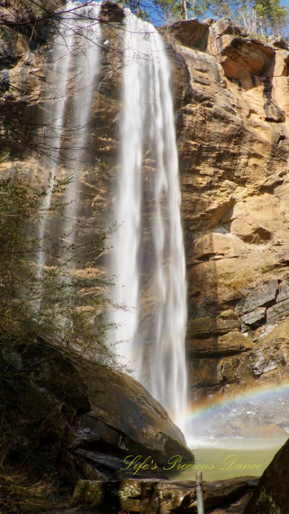 An 186 foot waterfall spilling over a rock face into a pool of water at Toccoa Falls. A rainbow is reflecting in the mist.