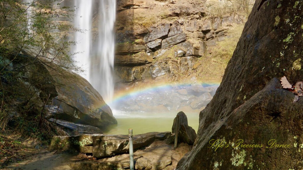 A rainbow reflecting in the mist above a pool of water as Toccoa Falls cascades down the rock face.